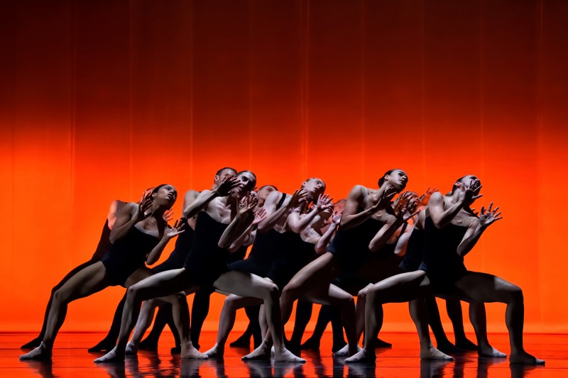 The dancers of Ballet Bc on stage with orange lighting. They are grouped together in a deep plie, wearing black bodysuits.