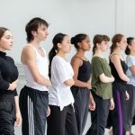 7 dancers stand in a diagonal line, each with their right hand crossed over their body.