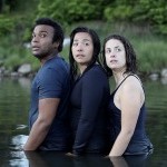 Three dancers stand close together, waist deep in the ocean with trees behind them, looking back with presence and hope