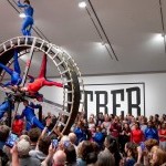 Dancers suspend in a giant wheel with large audience surrounding them