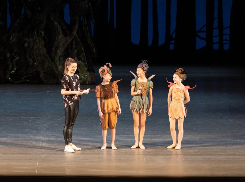 Principal dancer Megan Fairchild holds a microphone and speaks with four students, who are dressed in bug costumes.