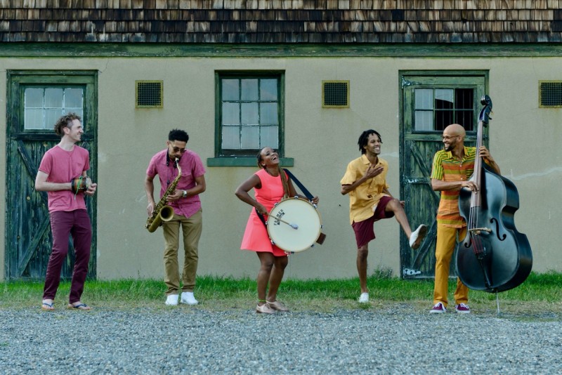 Music From The Sole dancers musicians wearing bright colors jam in a line in front of a farmhouse building