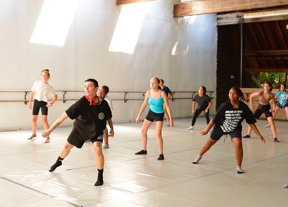 Sunlight falls through sky lights while a dance class takes place in a high-ceiling studio with a marley floor.