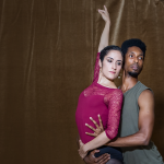 Two ballet dancers gaze past the camera in an embrace with arms extended 