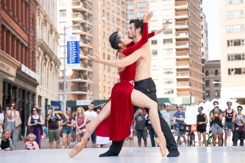 Two ballet dancers embrace on an outdoor city stage. 