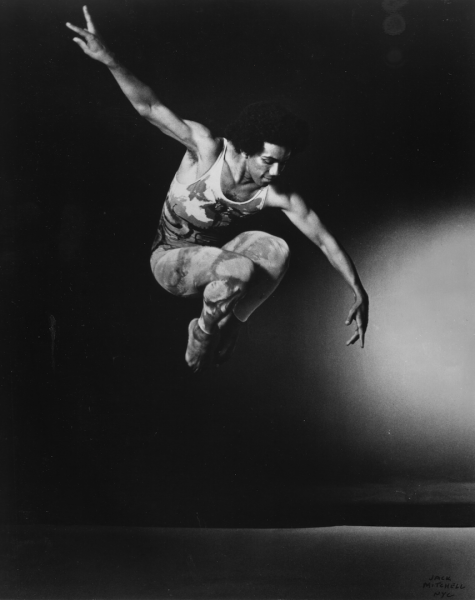 A Black dancer jumps in the air with his knees bent into his chest and his arms outstretched diagonally.