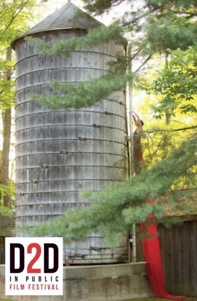 A blonde haired woman climbs a water tower with a long red dress trailing below her 