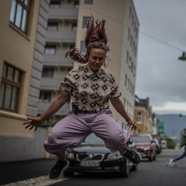 A woman with reddish-brown hair jumps mid-air on a city street, her hair flying upwards. She wears a patterned shirt and lavende