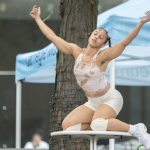 Dominica Greene is the dancer featured in this photo, wearing shorts and a tank top, with arms stretched out, kneeling on a plat