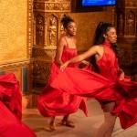 Two dancers with brown skin and natural hair dance wearing swirling red dresses