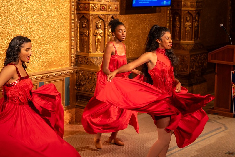 Two dancers with brown skin and natural hair dance wearing swirling red dresses