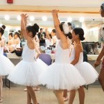 several ballerinas of various skin colors rehearsing in white tutus in front of a dressing room mirror with their instructor