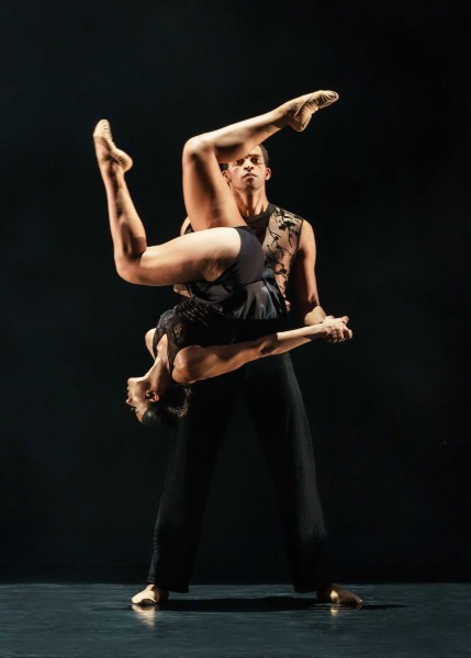 Two Philadanco dancers against a black background. One is in mid-air performing a flip while the other dance spots them.