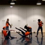 Dancers in various poses, various levels, wearing orange tops, inside a dance studio