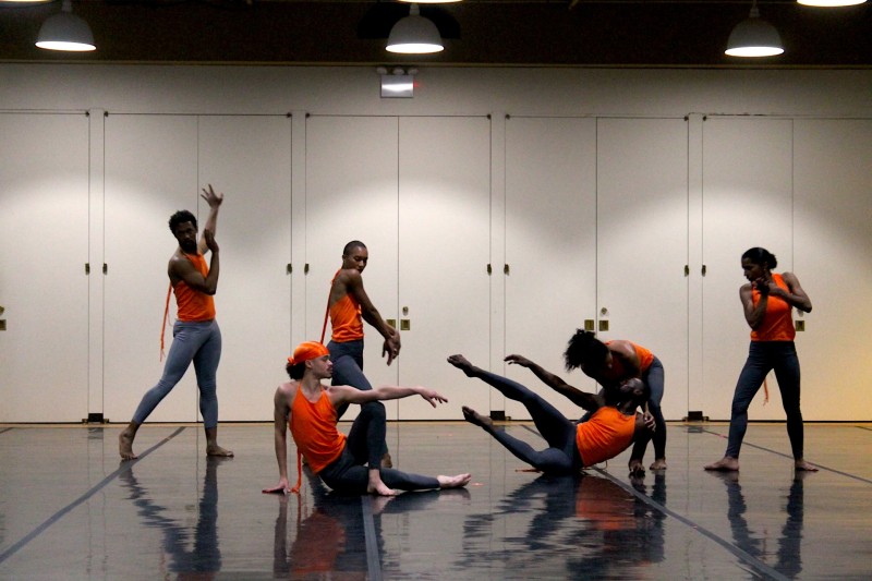 Dancers in various poses, various levels, wearing orange tops, inside a dance studio