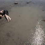 a woman lays, curled loosely around herself, in a thin layer of water on a grey mud flat. 