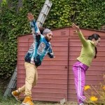 two dancers pictured from the side, jumping with arms outstretchend in. a community garden