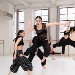 Dancer in black outfit jumping off other dancer, facing forward