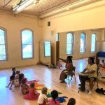 Kids sitting on the floor with rainbow scarves watching teacher talk