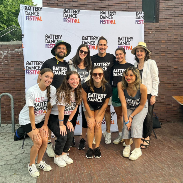 A group of volunteers wear Battery Dance t-shirts and stand in front of a Battery Dance Festival step and repeat.