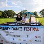 Three women sit at a table in an outdoor park selling Battery Dance merchandise