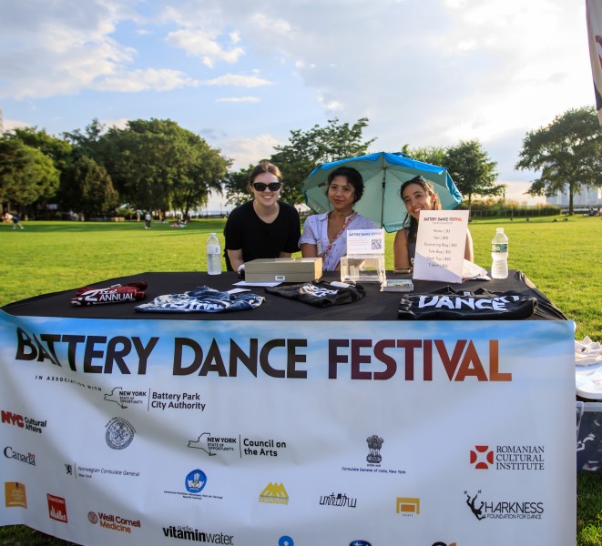 Three women sit at a table in an outdoor park selling Battery Dance merchandise