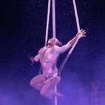 Young beaming Black aerialist performs on a trapeze in the rain with diamond print costume and dark purple lighting.