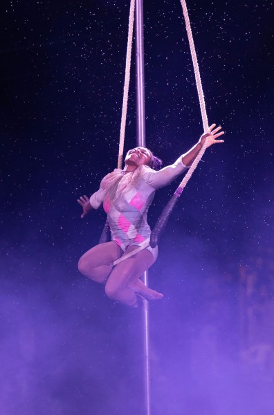 Young beaming Black aerialist performs on a trapeze in the rain with diamond print costume and dark purple lighting.