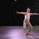Dancer stands in first plie with arms stretched to sides and hands gesturing.