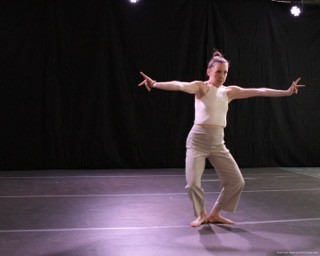 Dancer stands in first plie with arms stretched to sides and hands gesturing.
