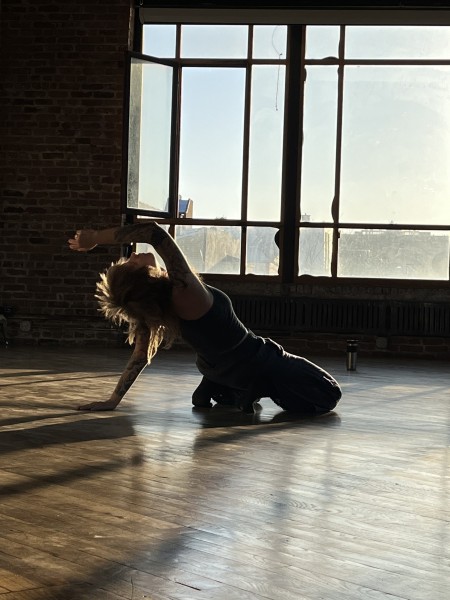 A person dances in a space with wooden floors, leaning back, the sun creating a shadow of their body.