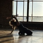 A person dances in a space with wooden floors, leaning back, the sun creating a shadow of their body.