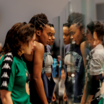 Two female identifying dancers looking at their reflection in a glass prop piece as part of a live art performance piece. 