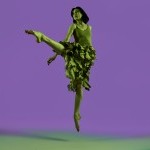 Dancer in green jumps in a crossed position resembling the letter "L" in front of a purple backround.