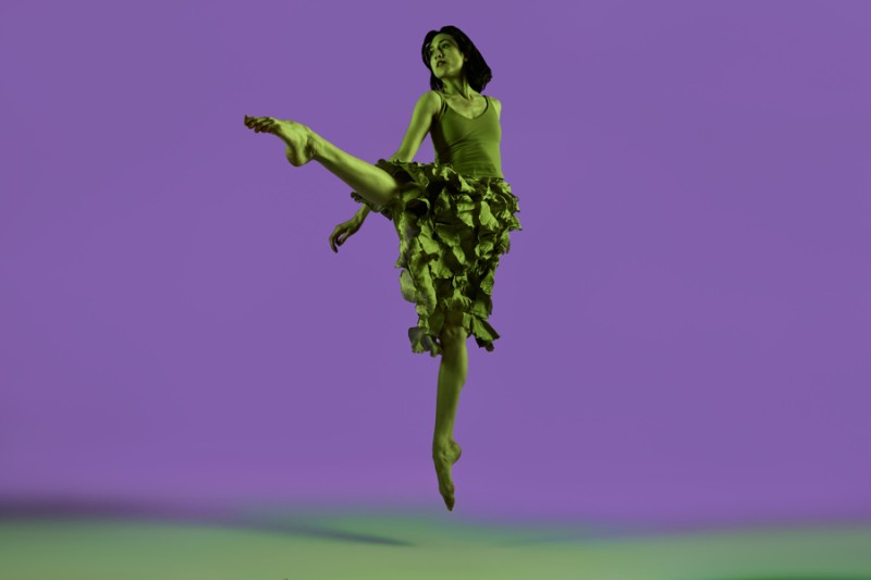 Dancer in green jumps in a crossed position resembling the letter "L" in front of a purple backround.