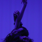 Two dancers lean on one another, back to back with their arms floating above them against and deep violet blue background. 