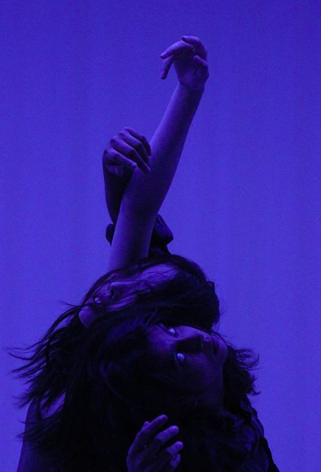 Two dancers lean on one another, back to back with their arms floating above them against and deep violet blue background. 