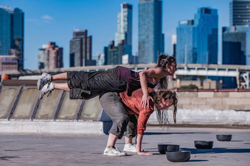 a woman in grey pants and purple top is balanced in a plank on the back of another dancer in an orange top city skyline behind