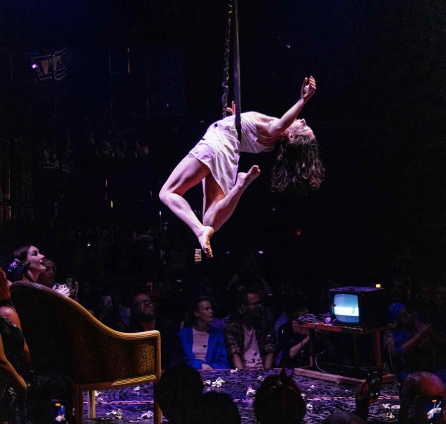  a dancer floats in the air surrounded by a vintage chair and little television with static on the screen. an audience looks on.
