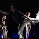 Dancer in foreground wearing white pants and a dark shirt pointing forward with one hand, back leg is in attitude.