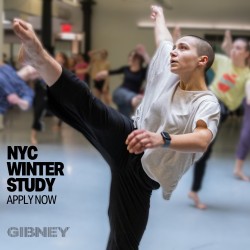 A dancer in a white t-shirt and black pants lifts their arm and extends their leg into the air