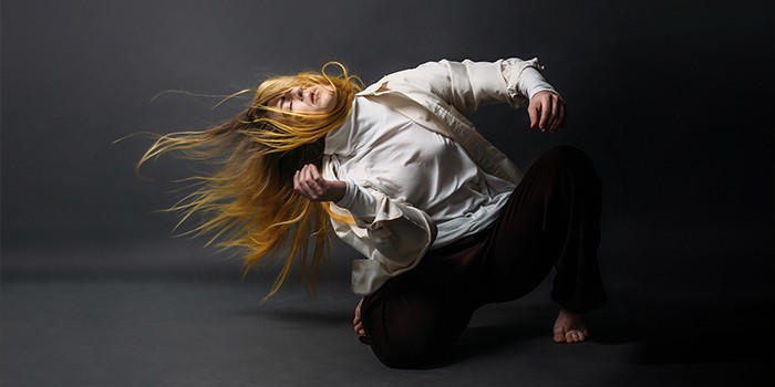 a female dancer with long blonde hair in a white shirt and brown pants, posing on her knees