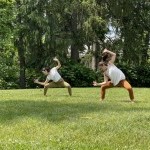 Two people dancing side by side in the grass in front of a line of trees.