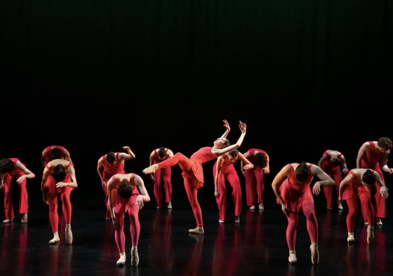 Group of dancers wearing pink costumes and contracted over. Solo dancer in the center of the stage is in a high release.