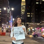 A lady holds a copy of the newspaper, Artistic Tribe NYC, while posing in the middle of the streets of Manhattan, at night. 