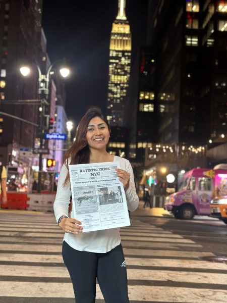 A lady holds a copy of the newspaper, Artistic Tribe NYC, while posing in the middle of the streets of Manhattan, at night. 