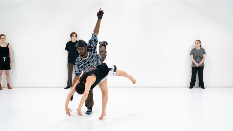 Two dancers create a partnering pose in front of three dancers watching