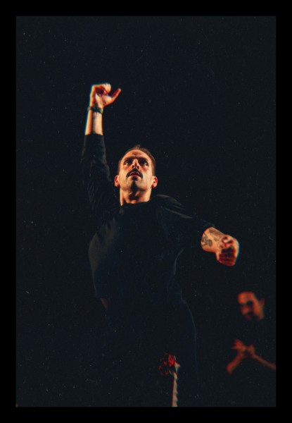 A man stands against a dark background dancing, while another man is in the background.