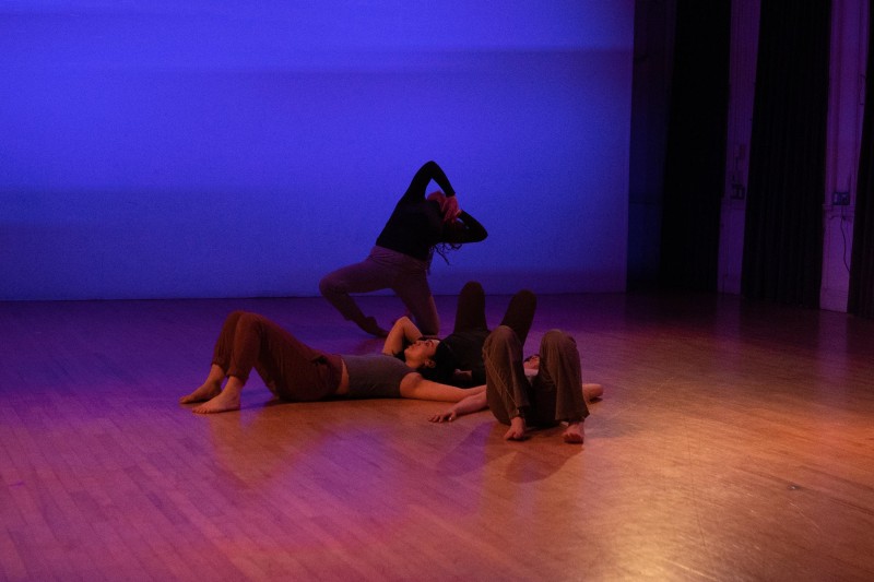 Three dancers lying on the ground connected while one dancer leans backwards on a medium level.