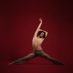 Male dancer in a straddle pose in front of red background with one arm up and shirtless.